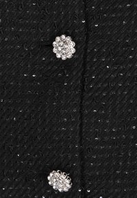 Juodas tekstūruotas audinys su zigzago raštu, papuoštas dviem gėlių formos sidabriniais mygtukais, dekoruotais skaidriais rhinestones.