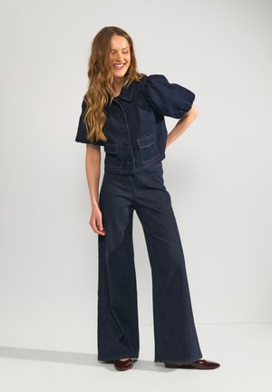 Femme aux longs cheveux ondulés portant une veste boutonnée bleu foncé à manches courtes et un jean à jambes larges, souriante, main sur la hanche.