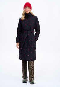 Čierny prešívaný dlhý kabát s červenými bodkami a opaskom, nosený cez nohavice s leopardím vzorom a zelenými snežkami, doplnený červenou čiapkou.