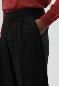 Zwarte op maat gemaakte broek met plooien, met een gladde textuur en een voorkantzak. Gecombineerd met een rood shirt met lange mouwen.