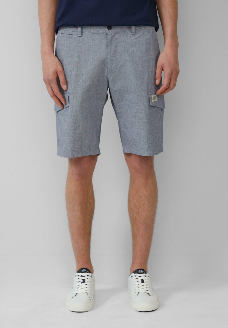 Pantaloni cargo a righe bianche e blu con tasche laterali, realizzati in un tessuto leggero. Indossati con scarpe da ginnastica bianche con dettagli blu navy.