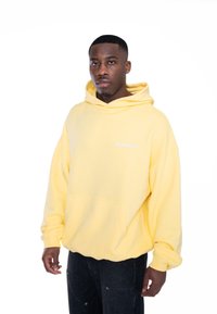 Sudadera amarilla con capucha y bolsillo frontal, con "MULTIPLY" impreso en blanco. Usada con pantalones oscuros, mostrando un ajuste relajado y una textura suave.