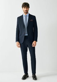 Marineblauer Anzug mit tailliertem Sakko und geraden Hosen. Hellblaues Hemd und gemusterte Krawatte. Schwarze Loafers und ein kastanienbraunes Einstecktuch.