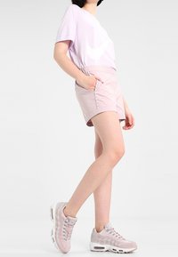 Ein helllila, übergroßes T-Shirt mit einem weißen Logo, kombiniert mit hellrosa Shorts mit seitlichen Taschen und einer Kordel. Dazu werden rosafarbene Sneakers getragen.