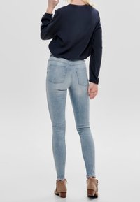 Jean skinny bleu clair avec deux poches arrière, associé à une chemise noire à manches longues. Le modèle porte des bottines marron, vu de dos.