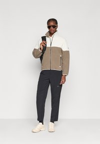 Giacca in pile di colore crema e marrone chiaro, con zip e colletto alto. Abbinata a pantaloni cargo scuri e sneakers beige chiare.