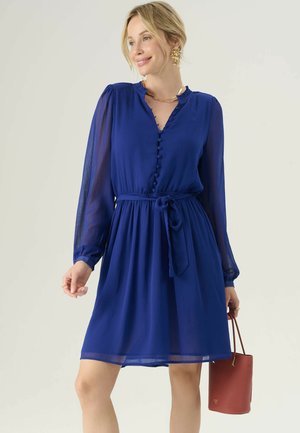Robe bleu royal avec de longues manches transparentes, décolleté en V, fermeture à boutons, ceinture réglable à la taille et jupe évasée, accompagnée d'un sac à main rouge.