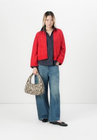 Vrouw staat in een rode jas, zwarte shirt, wijde spijkerbroek, zwarte flats, en houdt een tas met slangenhuidpatroon vast tegen een witte achtergrond.