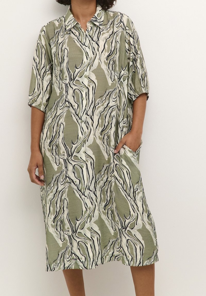 Personne portant une robe ample boutonnée longueur genou avec un motif abstrait ondulé vert, beige et noir et des manches trois-quarts, une main dans la poche.