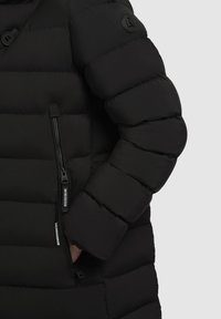 Close-up van een zwart gewatteerd jacksleeve met een hand in een ritszak en een klein, rond logo op de bovenarm.