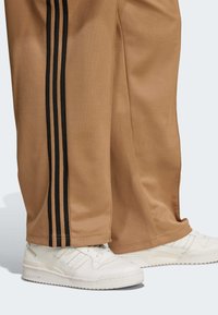 Pantalons de survêtement marron avec trois rayures noires sur les côtés, fabriqués en tissu doux. Associés à des baskets blanches avec des accents texturés.