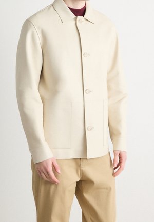 Mann trägt eine beige geknöpfte Jacke über einem dunkelroten Hemd und hellbraunen Hosen und steht vor einem einfarbigen weißen Hintergrund.