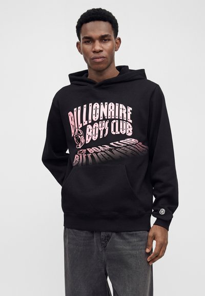 Νέος άνδρας με μαύρο φούτερ με ροζ κείμενο "Billionaire Boys Club" και γραφικό αστροναύτη, σε συνδυασμό με γκρι τζιν, μπροστά σε μονόχρωμο φόντο.