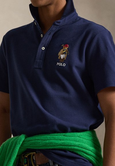 Μπλε ναυτικό πόλο μπλουζάκι με γιακά, στολισμένο με κεντημένη αρκούδα με κόκκινο καπέλο και κίτρινο παλτό. Πράσινο πουλόβερ δεμένο γύρω από τη μέση.