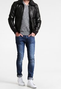 Černá kožená bunda s ovčím límcem, šedý svetr s výstřihem do V, modré úzké džíny a bílé tenisky. Hladké textury a přiléhavý design.