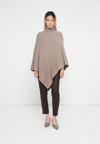 Poncho de lã castanho com colarinho alto, apresentando uma forma triangular, textura suave e mangas esvoaçantes, combinado com calças castanhas escuras e sapatos claros.