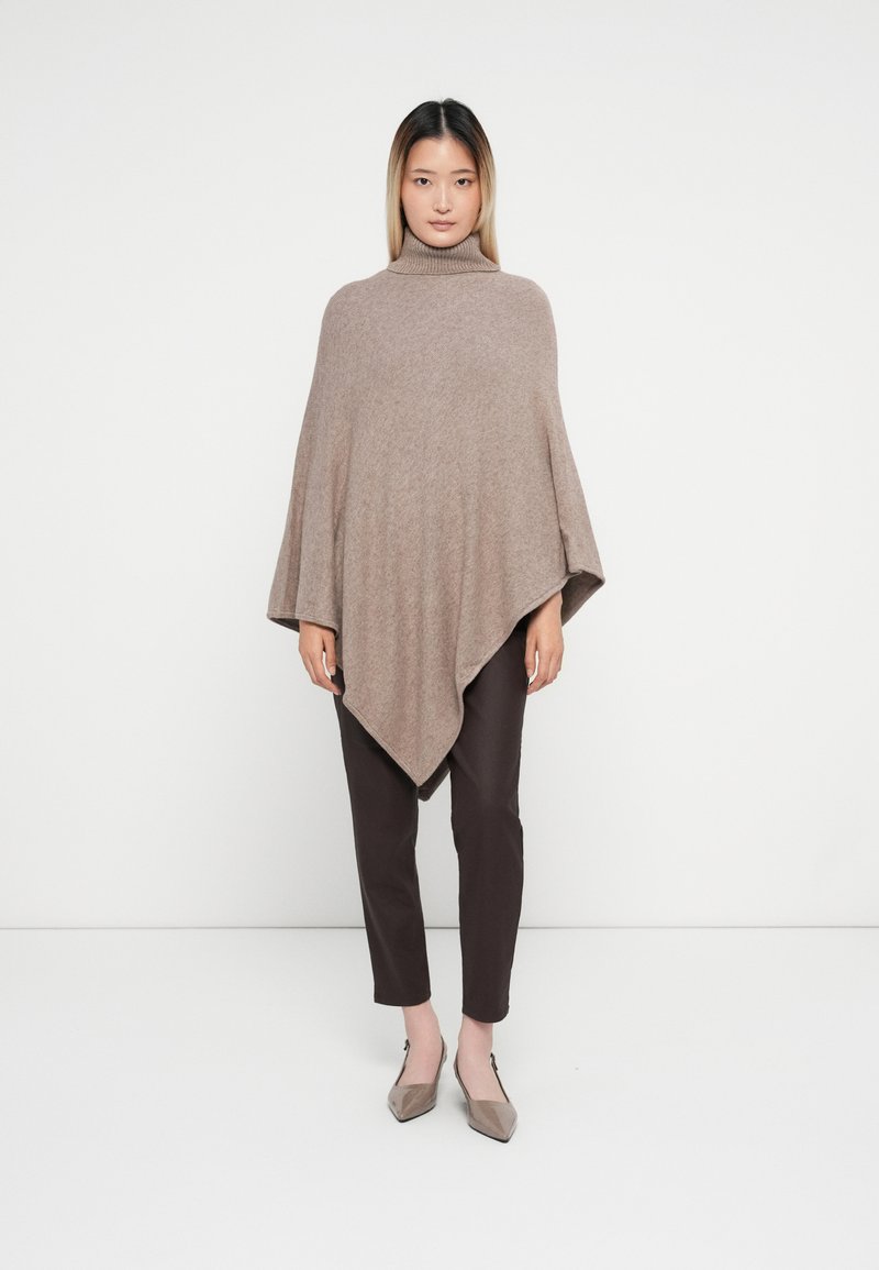Poncho de lã castanho com colarinho alto, apresentando uma forma triangular, textura suave e mangas esvoaçantes, combinado com calças castanhas escuras e sapatos claros.