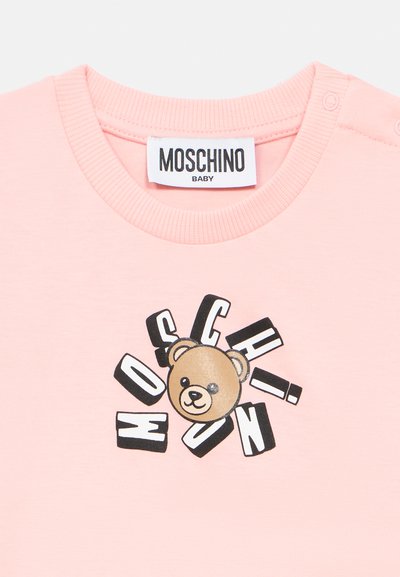 Ροζ ανοιχτό παιδικό πουκάμισο με ετικέτα της μάρκας Moschino, με το πρόσωπο μιας αρκουδίτσας περιτριγυρισμένο από σκορπισμένα ασπρόμαυρα γράμματα που σχηματίζουν τη λέξη "Moschino".