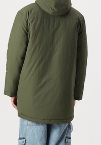 Chaqueta de plumas verde con capucha y corte relajado, que presenta una textura suave y una longitud más larga. Incluye una costura frontal y puños en las muñecas.