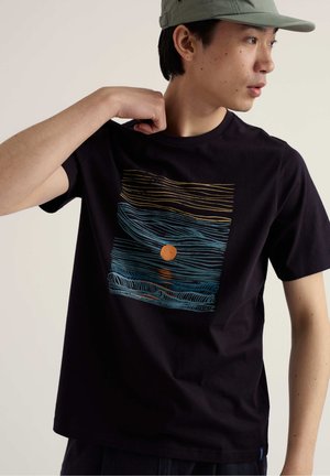 Uomo con berretto oliva e maglietta nera con design astratto di onde blu e arancioni e sole, che guarda verso destra, tirandosi il colletto della maglia.