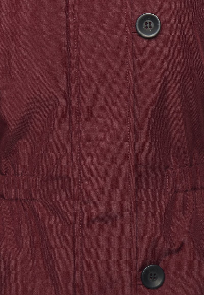 Veste isolante bordeaux avec une texture lisse, dotée d'une fermeture à boutons à l'avant et de détails élastiques sur les côtés.