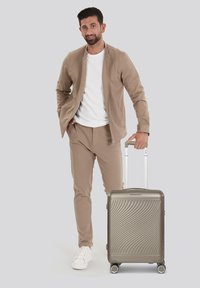 Zilveren harde koffer met golfpatroon, vier wielen en uittrekbare handgreep; model draagt een beige jas en broek met witte shirt en schoenen.
