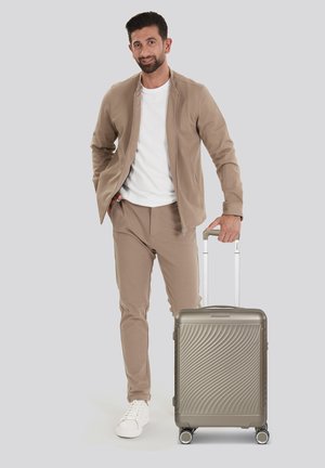 Silver hard-shell suitcase with wave pattern, four wheels, and retractable handle; model wearing beige jacket and pants with white shirt and shoes.