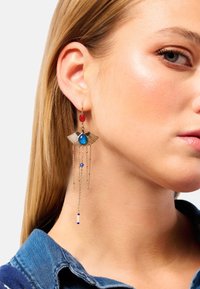 Boucles d'oreilles dorées ornées de pierres rouges et bleues, avec un design ailé, accompagnées de délicates chaînes et de petites perles, créant un effet superposé.