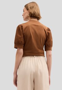 Braunes Cropped Top mit kurzen Puffärmeln und strukturierten Details, kombiniert mit hellbeigen, weit geschnittenen Hosen. Einfaches und figurbetontes Design.