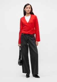 Červený svetr s texturovaným úpletem a dlouhými rukávy, s vázáním, v kombinaci s černými širokými džínami s vysokým pasem a černými kotníkovými botami.