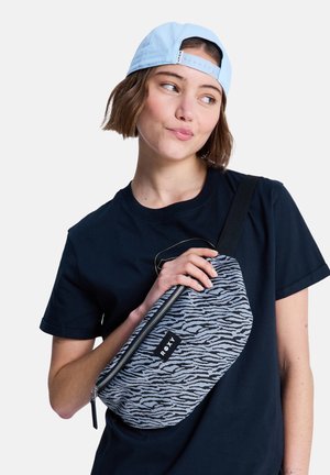 Junge Person mit einer hellblauen Kappe und schwarzem Hemd, die eine grau-schwarze Roxy-Gürteltasche mit Muster quer vor der Brust hält.