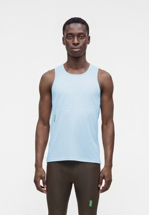 Homme aux cheveux courts portant une chemise sans manches bleu clair et un pantalon marron foncé ajusté, debout devant un fond clair uni.
