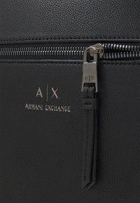 Fekete műbőr táska ezüst színű cipzárral, az "A|X ARMANI EXCHANGE" felirattal dombornyomva, és egy bőr díszítéssel ellátott cipzárhúzóval.