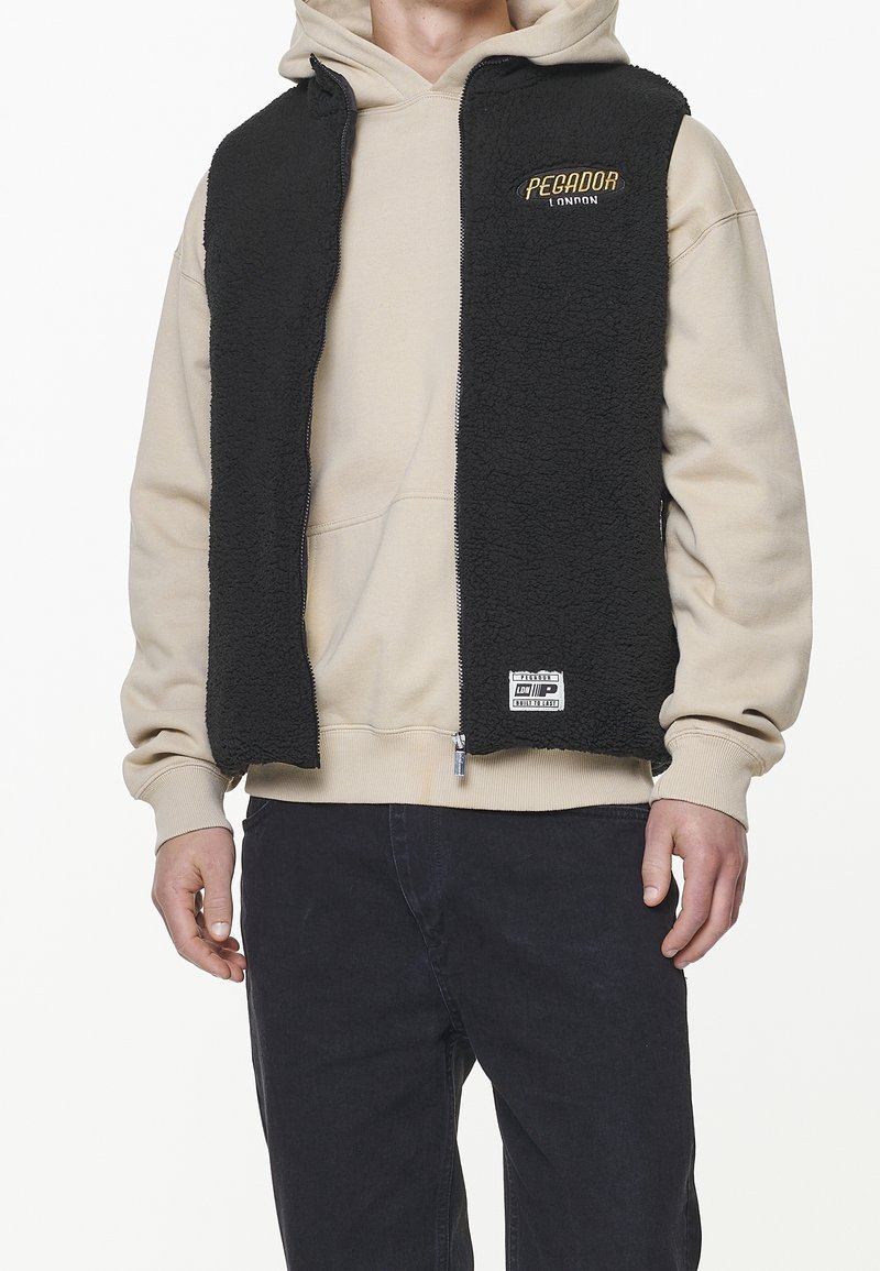 Svart fleeceväst med dragkedja framtill, dragen över en beige hoodie. Har en broderad logotyp och ett märke på nedre vänstra sidan.