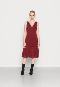 Vestido bordeaux com decote em V profundo e corpete justo; saia de chiffon esvoaçante na barra. Modelo usa botas pretas de tornozelo. Fundo liso.