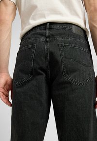 Man draagt zwarte Lee-jeans en wit overhemd, gezien van achteren van taille tot halverwege de dij, met zichtbare achterzakken en riemlussen.