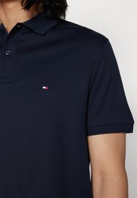 Marineblauwe polo gemaakt van katoen, met een knoopsluiting bij de kraag, korte mouwen en een klein rood-wit logo op de linkerkant van de borst.