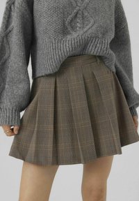 Bruine geruite rok met plooien en een gladde textuur, gecombineerd met een grijze gebreide trui. Zichtbare hand die de zoom van de rok vasthoudt.