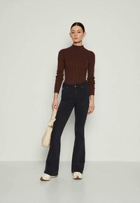 Suéter bege com gola alta canelada combinado com jeans flare azul escuro; modelo segura uma bolsa creme; fundo minimalista.