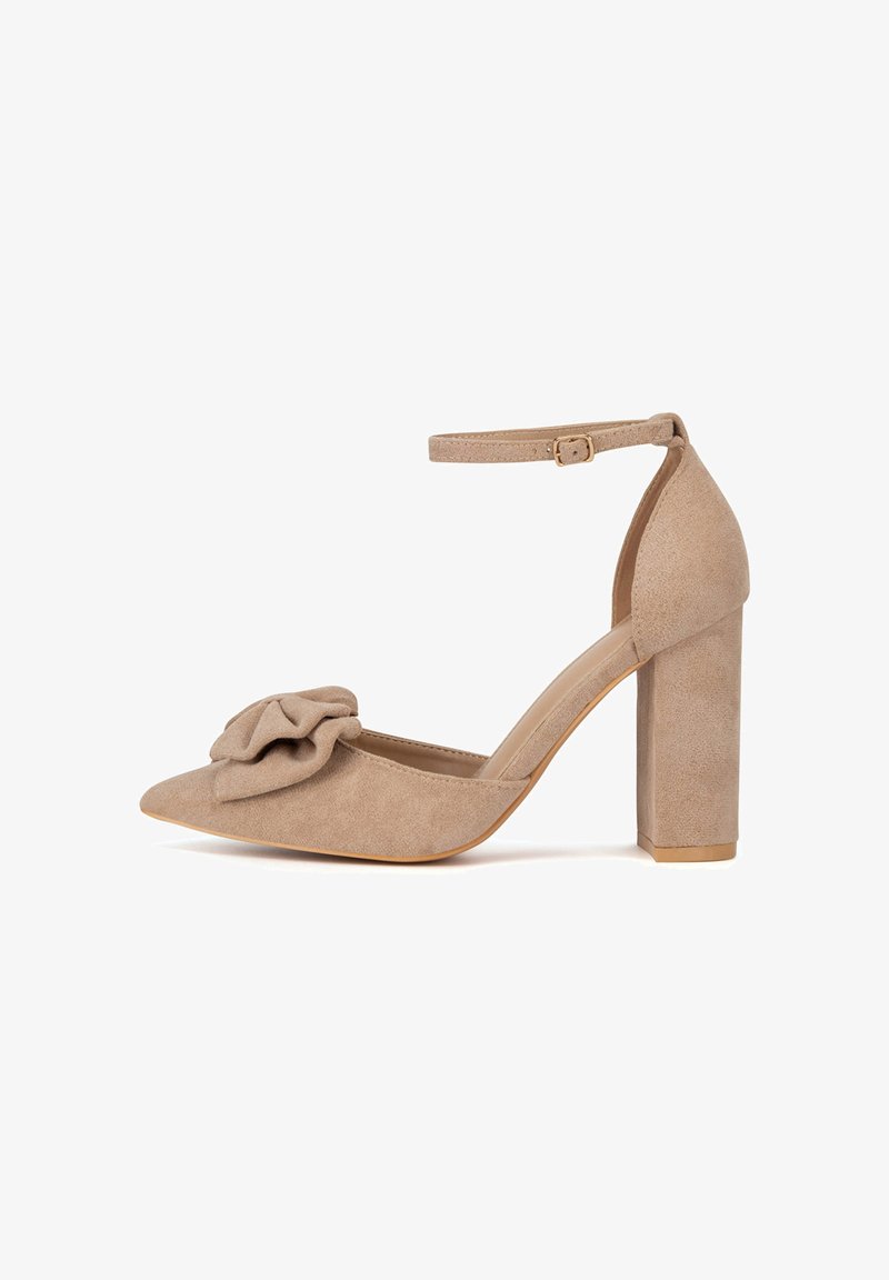 Zapato de tacón alto de ante beige con punta puntiaguda, tacón ancho de bloque y detalle decorativo de volantes en la parte frontal, asegurado con una correa en el tobillo.
