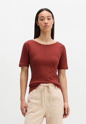 Femme aux longs cheveux foncés portant un t-shirt à manches courtes couleur rouille et un pantalon beige à cordon, debout devant un fond blanc uni.