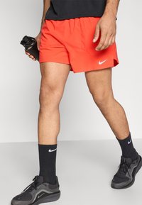 Κόκκινα σορτς Nike με ελαστική μέση, συνδυασμένα με μαύρες κάλτσες Nike και μαύρα αθλητικά παπούτσια, με ομαλή υφή και μίνιμαλ σχεδιασμό.