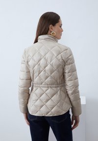 Donna con capelli castani lunghi che indossa una giacca imbottita beige e jeans scuri, vista di spalle su uno sfondo bianco uniforme.