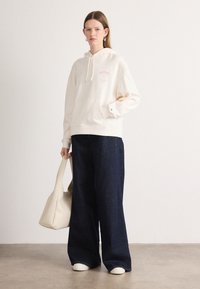 Felpa crema con tasca frontale, dettaglio grafico rosa, abbinata a jeans larghi scuri e scarpe bianche senza lacci; tiene una borsa tote beige.