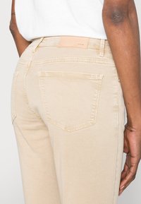 Vaqueros de mezclilla beige con una textura suave, bolsillos traseros y una etiqueta de cuero con "Opus" en la cinturilla. Diseño ajustado y pierna recta.