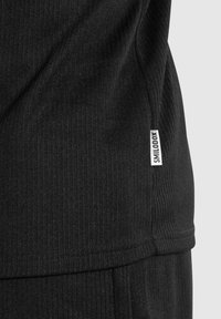 Schwarzer, gerippter Stoff, enganliegendes Design, mit einem kleinen weißen Label mit der Aufschrift "SMILODOX" an der Seitennaht. Strukturierte Oberfläche sichtbar.