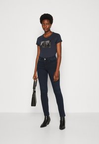 Marine t-skjorte med et mønstrede grått og svart design, kombinert med mørke skinny jeans og svarte ankelstøvler, holder en svart håndveske.