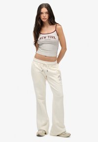 Joven mujer vistiendo una camiseta de tirantes gris con el texto "New York", pantalones de chándal anchos crema y zapatillas beige frente a un fondo blanco liso.