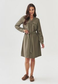 Olive green dress with long sleeves, front button closure, two chest pockets, and a tied belt at the waist. Soft, smooth fabric texture.
