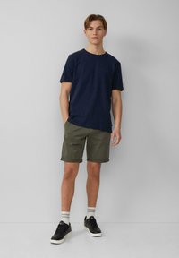 Temno plavi kratki rukav T-shirt u kombinaciji sa maslinastozelenim šortsevima s preklopljenim manžetama. Crne tenisice s bijelim potplatima i prugaste čarape upotpunjuju odijelo.